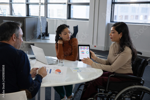 Diverse team reviewing reports at table in office, wheelchair woman presenting tablet, copy space