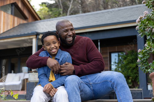 African American dad and child sitting on porch steps, smiling in denim jacket and burgundy sweater