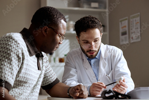 Tableau sur toile Black middle aged man consulting Caucasian male doctor, reviewing medical device during clinic visit