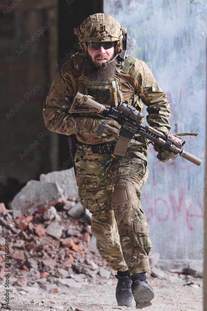 Fototapeta premium Bearded Soldier In Tactical Gear Running With Rifle Through Urban Ruins During Military Operation