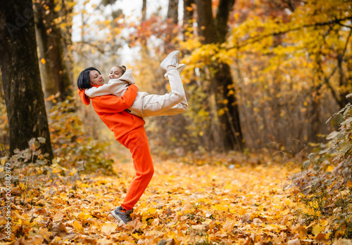 Mother embracing her little daughter among falling autumn leaves, authentic family bonding, sincere love and warmth in fall park	
