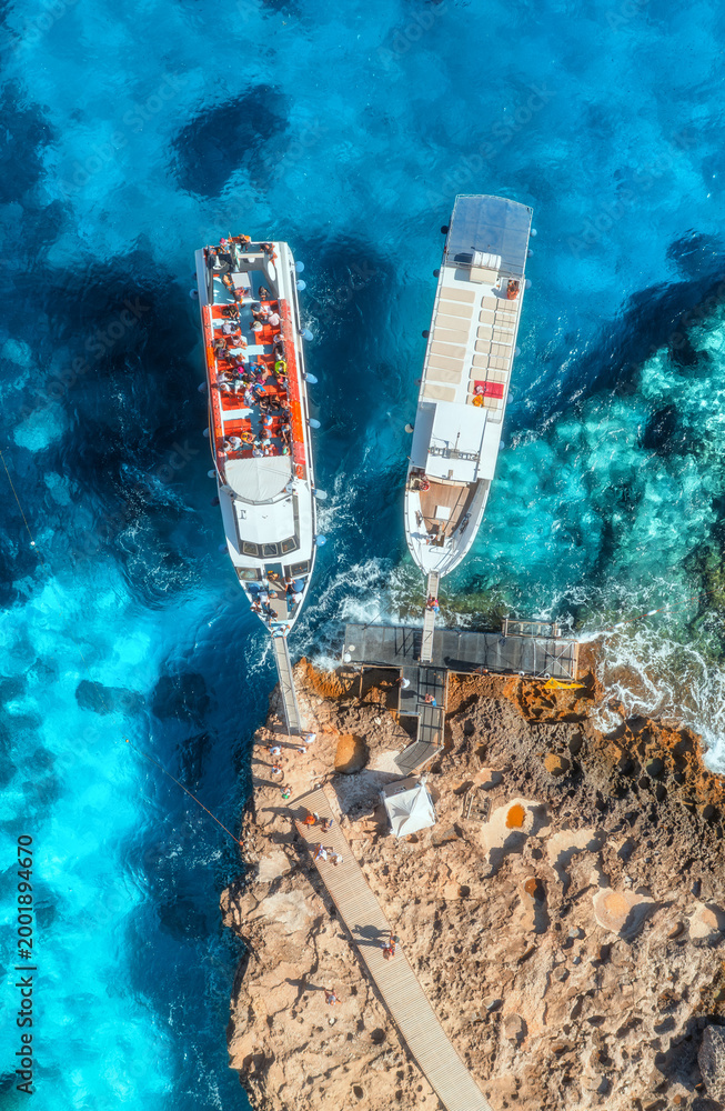 Fototapeta premium Aerial view of boat with people and rocky sea coast on sunny summer day. Top drone view of yacht, transparent azure water, path. Mediterranean sea. Travel in Sardinia island, Italy. Tropical landscape