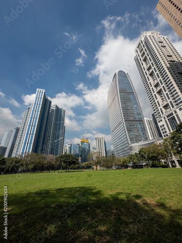 Quadro su tela Vibrant modern cityscape of Bonifacio Global City, Manila, showcasing impressive skyscrapers towering over a serene expanse of verdant parkland under a bright, partly cloudy day