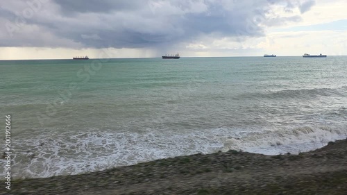 Moving shot from a train window viewing the turbulent sea with several cargo ships on the horizon under a dramatic, cloudy sky, while waves crash against the shore in the foreground