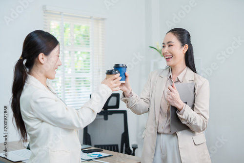 Two professional businesswomen are working on laptop computers sitting at desks in the office. Colleagues are happy and discussing the digital strategy of the Graph