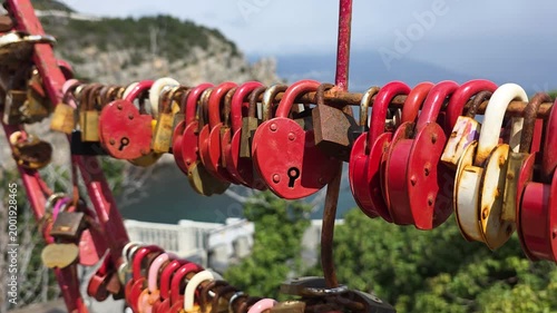 Red and rusted heart-shaped love padlocks symbolize everlasting commitment and romance, securely fastened to a bridge railing with an ocean vista in the background