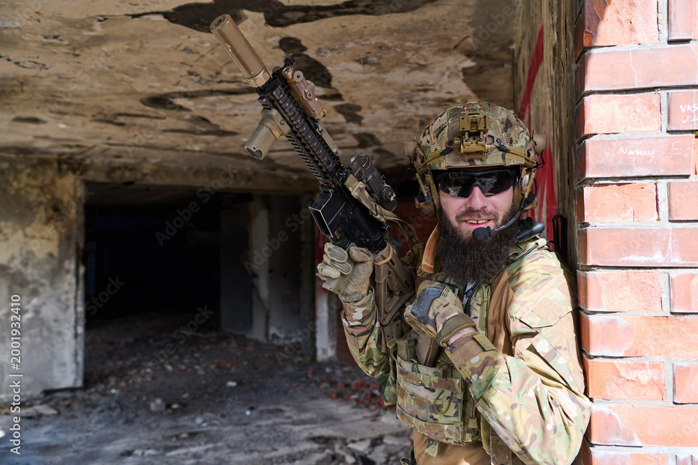 Fototapeta premium Bearded Soldier in Tactical Gear With Rifle Smiling Against Brick Wall in Urban Ruins