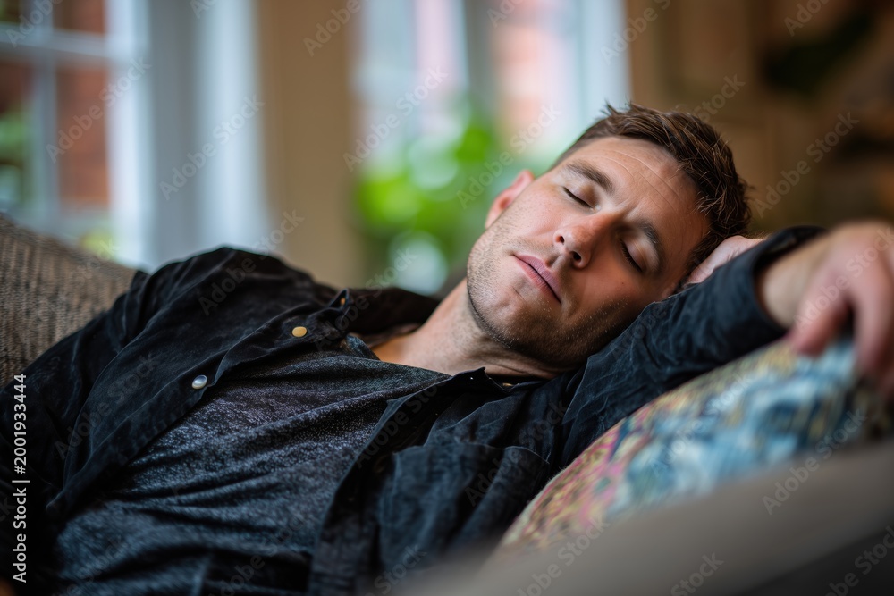 Fototapeta premium Man napping peacefully on a comfortable couch indoors, eyes closed, relaxed and resting. Head resting on arm