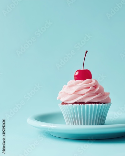Sweet cupcake with pastel pink frosting and bright red cherry on top, served on a matching light blue plate against a solid blue background. Minimalist aesthetic