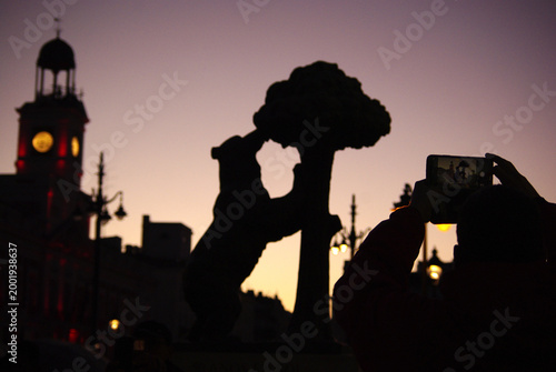 Unas manos y un teléfono móvil hacen una foto a las siluetas características de la ciudad de Madrid con la estatua del Oso de Madroño y el reloj de Puerta del Sol. Madrid