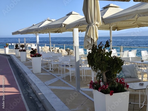 Картината върху платно Seaside dining terrace: white wicker and wooden chairs, cushioned seats with floral pillows, large cream umbrellas, and unobstructed blue‑sea views — airy, elegant Mediterranean dining by the water