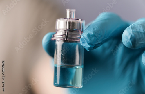 Photos Close-up of a hand in a medical glove holding a small vial with blue liquid, ide