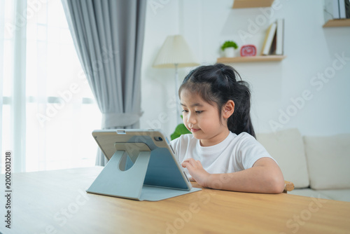 Young girl sitting at a wooden table engaging with a tablet device in a bright and cozy room with modern decor and natural light streaming through the window