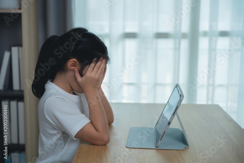 Young girl feeling frustration and sadness while looking at a tablet device on a wooden desk in a well-lit room with soft curtains in the background