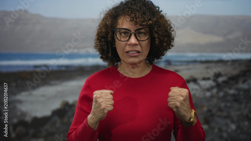 Photography Woman in bright red sweater clenches fists on rocky beach as waves crash behind; determination resilience