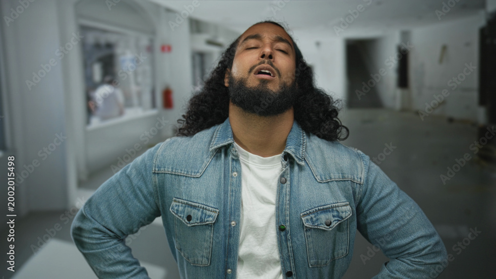Obraz premium Man with beard and long hair clutching chest and closing eyes in empty studio space, denim jacket visible, hand pressed to sternum, possible heartattack; pain vulnerability struggle recovery.