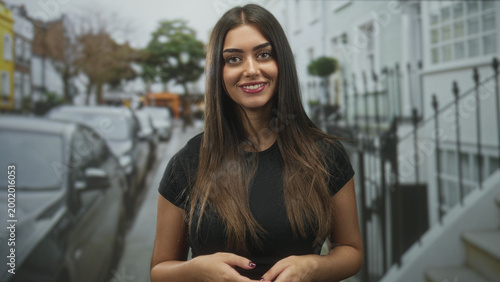 Foto Woman smiling, hands clasped at waist, facing camera on a residential street with parked cars and iron railings; confidence