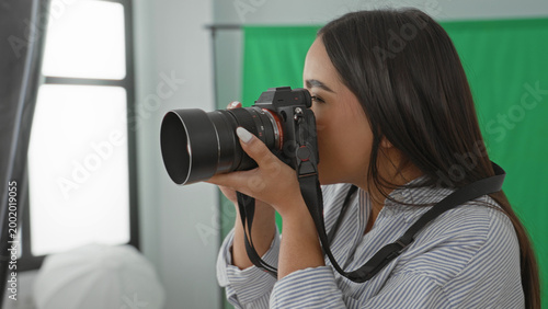 Foto Woman holding camera with hands on lens, looking through viewfinder and later checking playback while smiling in studio with green screen and softbox; creative joy