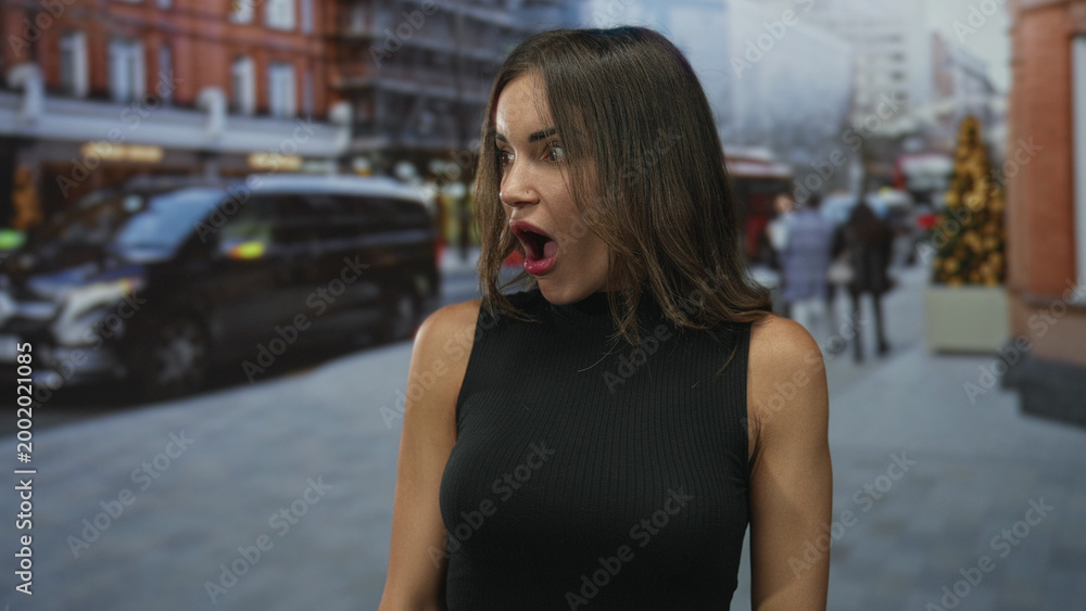 Fototapeta premium Woman in black sleeveless top with open mouth amid blurred pedestrians on street lined with vehicles and brick buildings; surprise reaction.