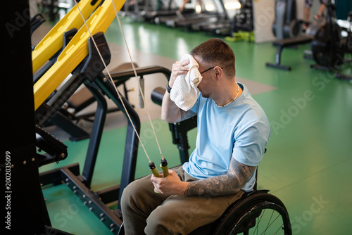фотографія Strong focused disabled man in wheelchair wiping sweat with towel while finishing workout on gym equipment at rehabilitation training