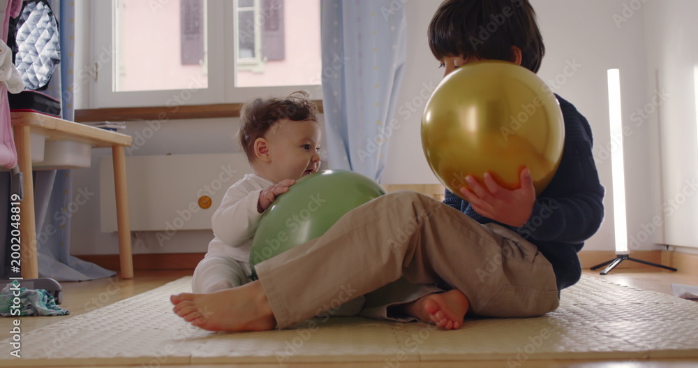 Fototapeta premium Baby and young boy playing together on floor with golden and green balloons in cozy bedroom, joyful sibling interaction and tender family bonding moment