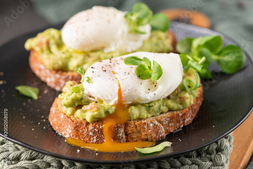 A beautiful close-up of two poached eggs on toasted sourdough bread with mashed guacamole and a vibrant runny yolk dripping down the side of the crusty bread on a black plate