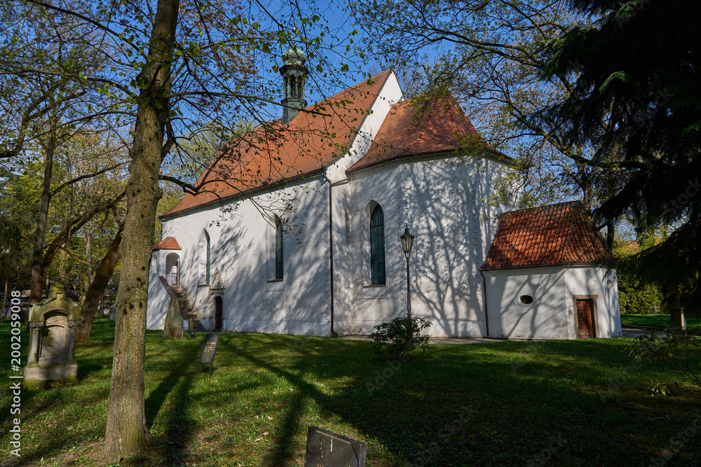 Fototapeta premium Pisek, Czech Republic - April 18, 2026 - Memorial park at the Holy Trinity. Historic white church with a red tiled roof surrounded by trees on a sunny day