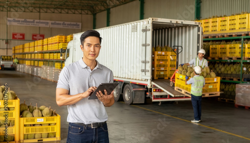 A confident logistics manager uses a tablet to oversee durian shipments while workers load crates into a container truck at a large-scale fruit distribution warehouse.