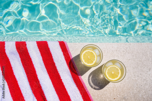 Two glasses of lemon drink with ice near swimming pool and red striped towel on sunlit terrace, summer lifestyle flat lay with copy space. Vacation relaxation, refreshing beverage and sunny poolside l