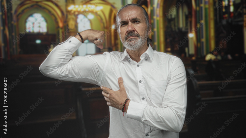 Obraz premium Man flexing bicep and pressing palm to chest inside ornate church building wearing white shirt and bracelets; faith confidence.