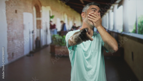 Foto Man pointing finger and covering mouth with hand in a corridor of a historic building, visible archways and columns; embarrassment apology regret