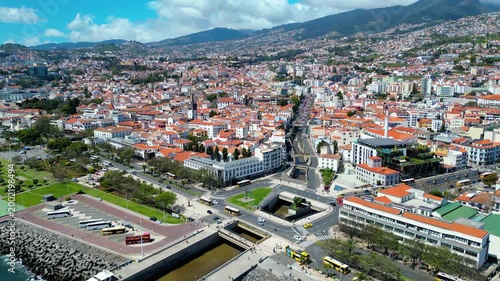 An expansive aerial view of Funchal, Madeira, showcasing its vibrant urban landscape nestled against lush green mountains under a bright blue sky. A bustling hub of culture and beauty.
