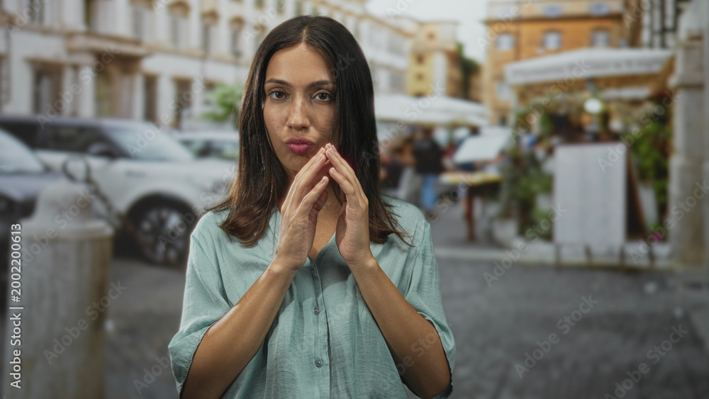 Fototapeta premium Hispanic brunette young woman blowing a kiss with both hands near mouth on a cobbled street in town; playful flirtation.