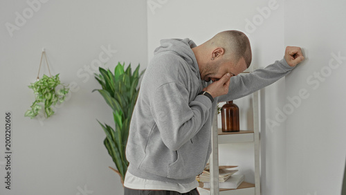 Man in gray hoodie leans on shelving unit, holds frame edge and rubs eyes in building; fatigue frustration reflection.