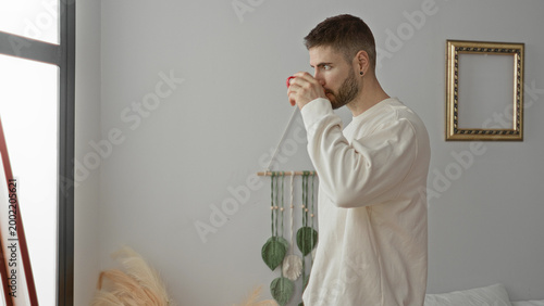 Tableau sur toile Man holding red mug and sipping coffee beside bed near a window in a bedroom; morning routine serenity