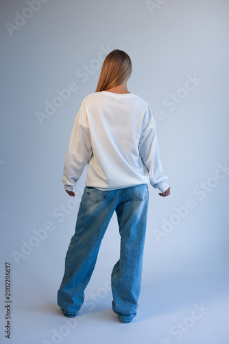 Beautiful Young Blonde Woman Standing Back View in Oversized White Blank T-Shirt Mockup and Blue Jeans on Light Studio Background
