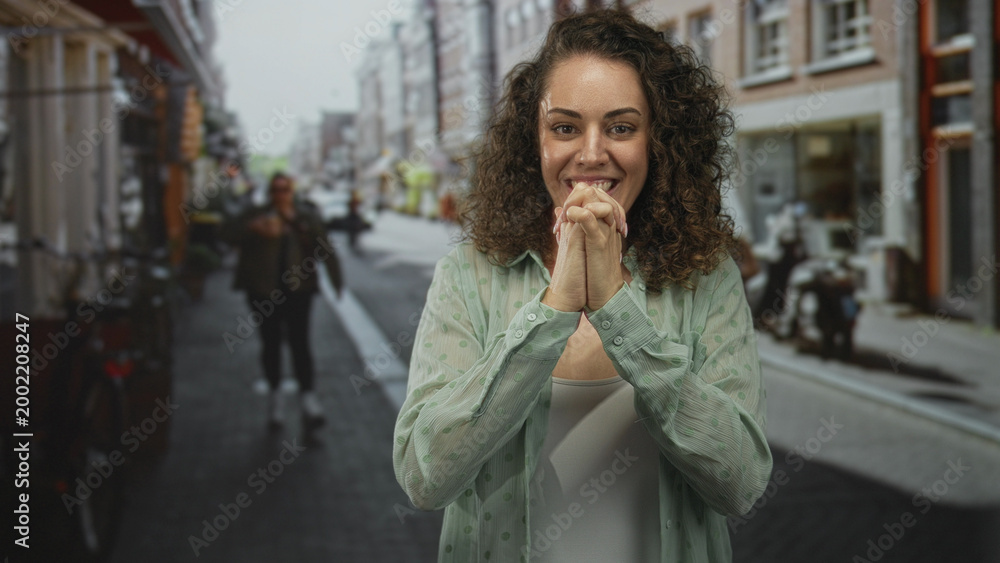 Fototapeta premium Young hispanic woman smiling with fingers crossed and hands pressed together in prayer on a city street lined with shops; hopeful.