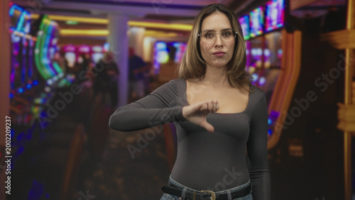 Foto Woman wearing glasses gives thumbs down gesture showing chest and stern expression amid slot machines in a casino building; disapproval loss