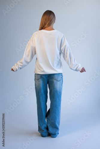 Beautiful Young Blonde Woman Standing Back View in Oversized White Blank T-Shirt Mockup and Blue Jeans on Light Studio Background