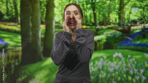 Εκτύπωση καμβά Beautiful hispanic woman cupping hands around mouth shouting in green forest park by pond and flowerbed, wearing dark long sleeve top; urgent call