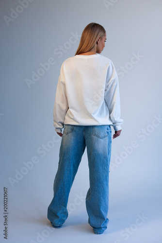 Beautiful Young Blonde Woman Standing Back View in Oversized White Blank T-Shirt Mockup and Blue Jeans on Light Studio Background