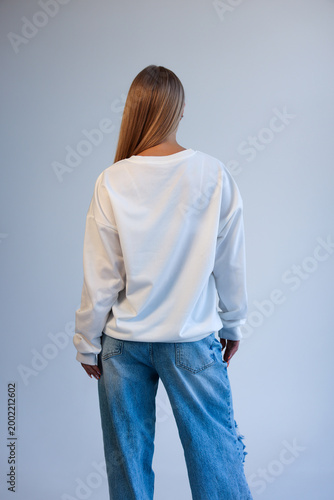 Beautiful Young Blonde Woman Standing Back View in Oversized White Blank T-Shirt Mockup and Blue Jeans on Light Studio Background