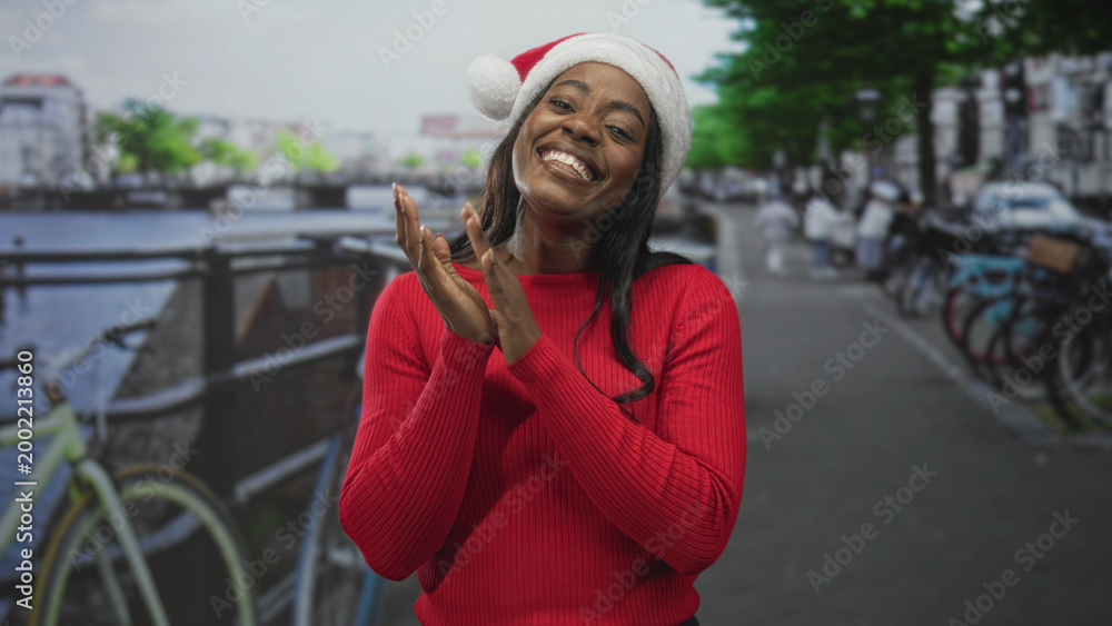 Naklejka premium Woman wearing santa hat and red sweater smiling and clapping hands on a canal street lined with bicycles and railing; holiday joy warmth.