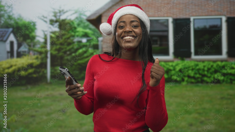 Fototapeta premium Woman in santa hat holding smartphone and giving thumbs up in front of a building, smiling and dancing subtly; holiday cheer joyful.