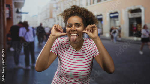 Foto Young african american woman in a striped t shirt pulls her cheeks and sticks out her tongue while making playful faces on a crowded city street; playful expression
