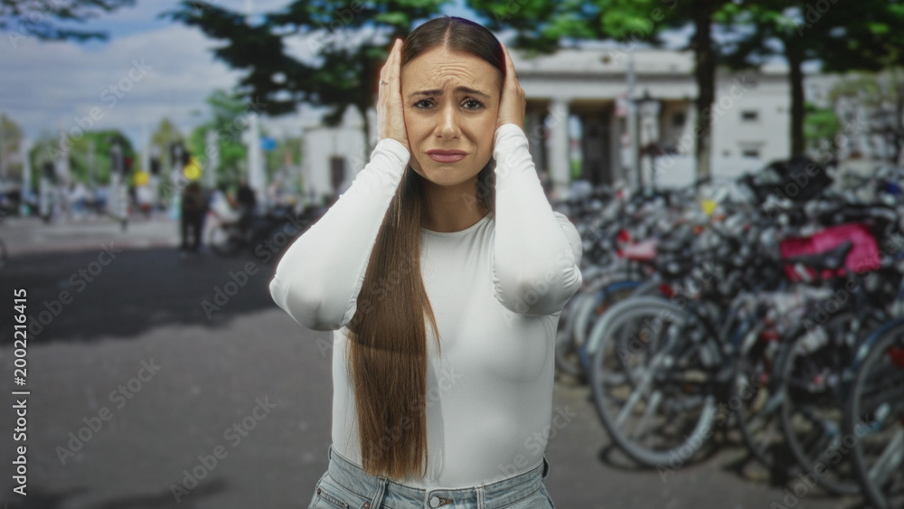 Fototapeta premium Woman covering ears with hands beside crowded bicycle rack on busy city street near columns and trees; urban stress.
