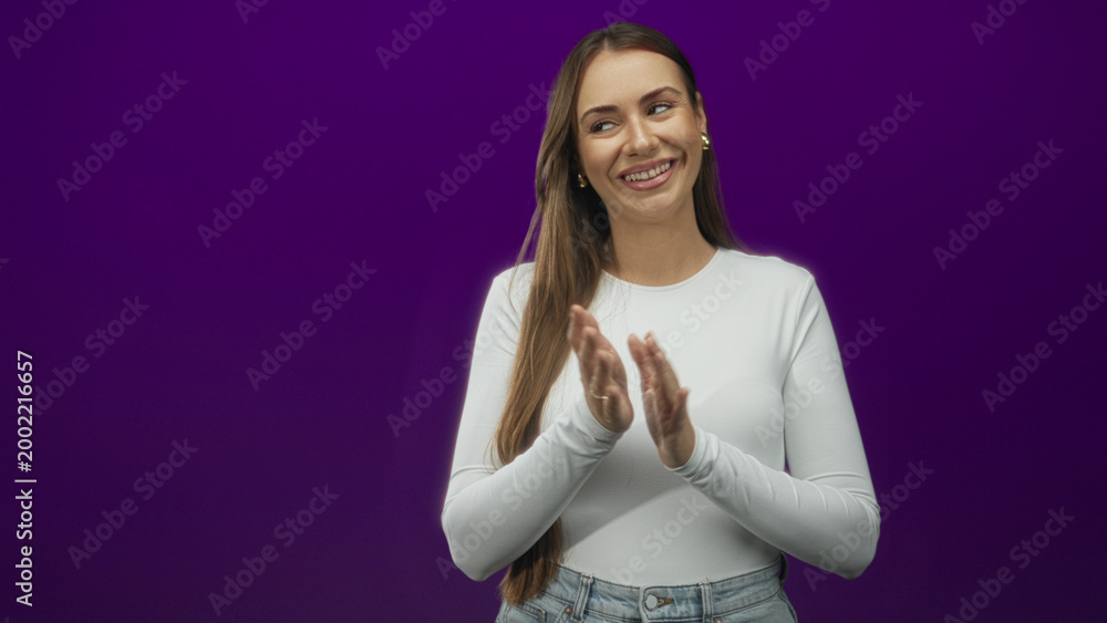 Fototapeta premium Young hispanic woman clapping hands with closed eyes and soft smile in a purple studio setting, wearing white shirt and jeans; joy celebration.