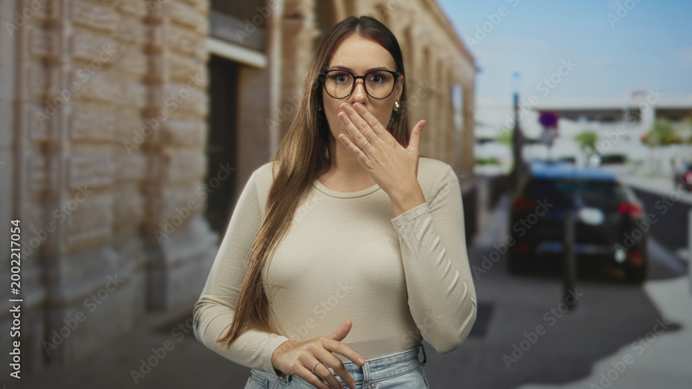 Fototapeta premium Young woman covers mouth with both hands, wearing glasses and jeans while standing by a stone building and parked car on street; surprise secrecy.