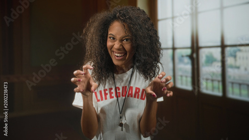 Young woman lifeguard tshirt with whistle on lanyard, hands splayed reaching toward camera in a building; playful confidence.