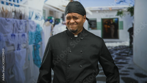 Photos Hispanic man chef in black uniform grimaces, face scrunched while standing in a narrow street lined with laundry; discomfort resilience